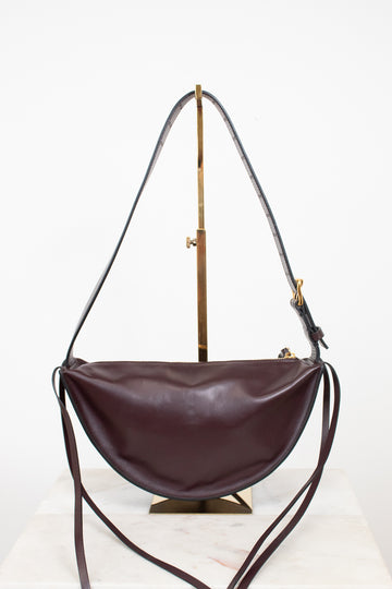 A dark burgundy leather crescent-shaped shoulder bag with a long thin strap, displayed on a gold-colored stand against a white background.