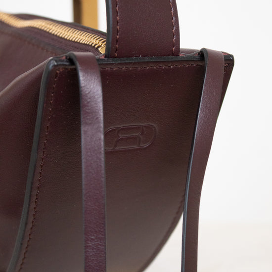 Close-up of a dark burgundy leather handbag with a gold zipper and a debossed logo on the side, set against a clean white background.
