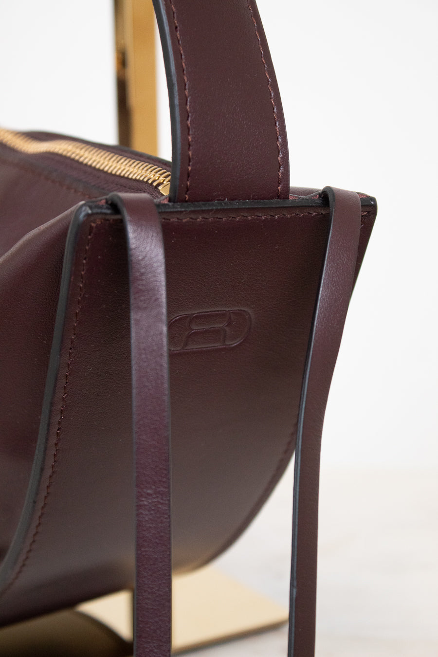 Close-up of a dark burgundy leather handbag with a gold zipper and a debossed logo on the side, set against a clean white background.