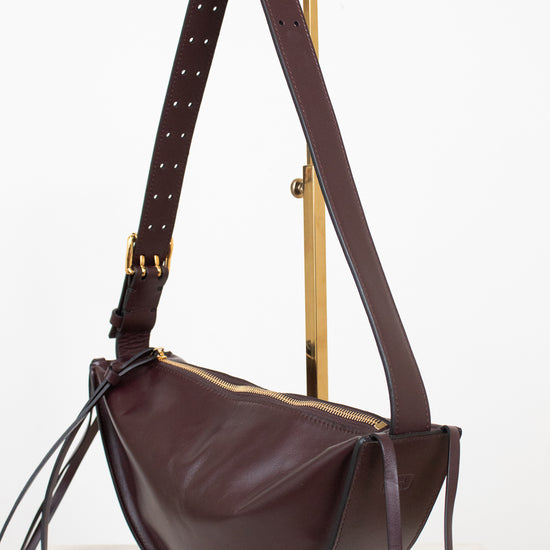 A dark burgundy leather crescent-shaped shoulder bag with gold-tone hardware, displayed on a gold metal stand against a clean white background.