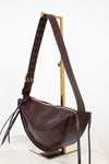 A dark burgundy leather crescent-shaped shoulder bag with gold-tone hardware, displayed on a gold metal stand against a clean white background.