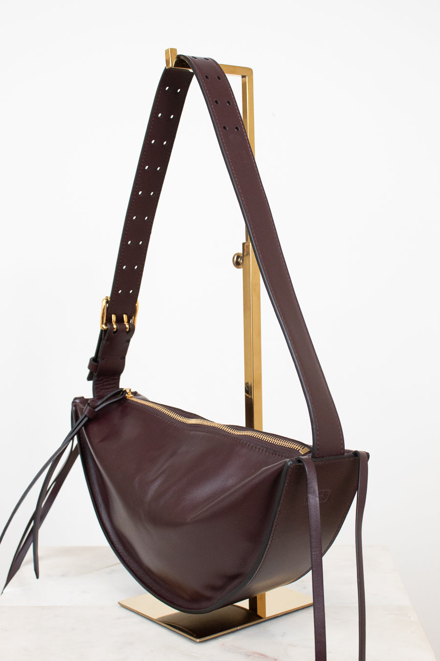 A dark burgundy leather crescent-shaped shoulder bag with gold-tone hardware, displayed on a gold metal stand against a clean white background.