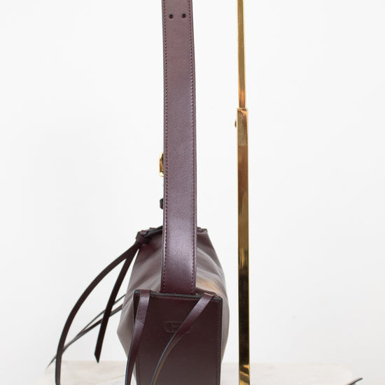 A dark burgundy cresent bag with a long matching shoulder strap, displayed on a gold metal stand against a clean white background.