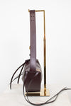 A dark burgundy cresent bag with a long matching shoulder strap, displayed on a gold metal stand against a clean white background.