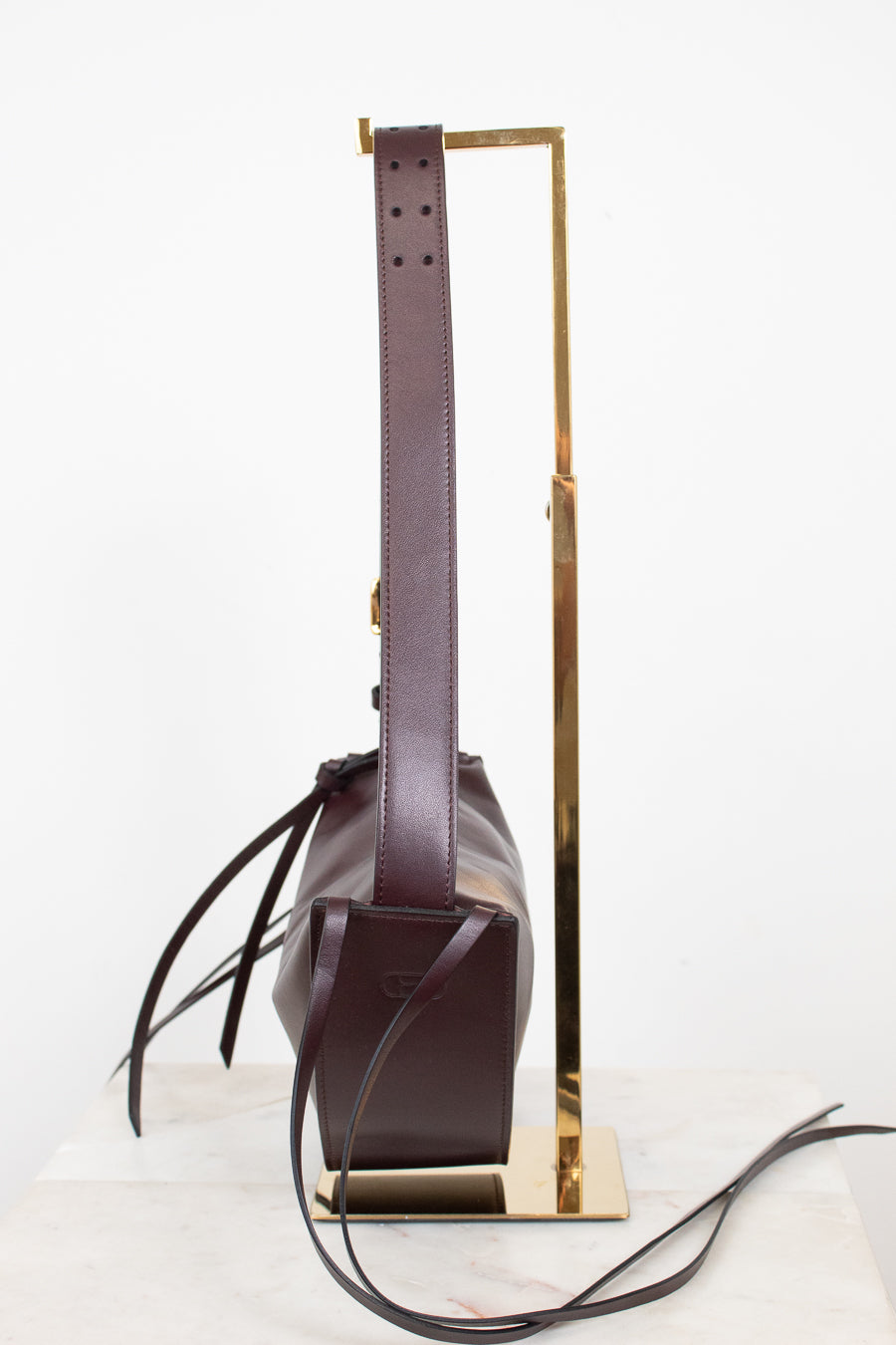 A dark burgundy cresent bag with a long matching shoulder strap, displayed on a gold metal stand against a clean white background.