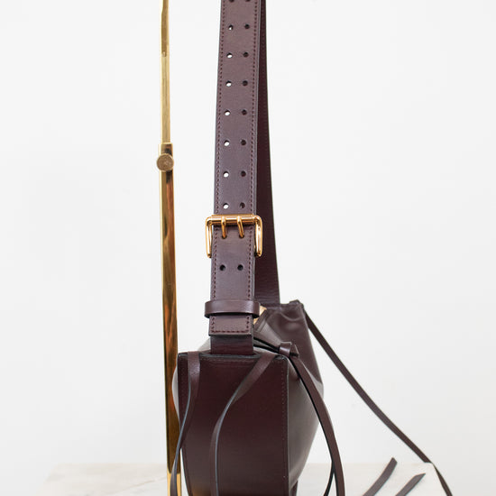 A dark burgundy leather shoulder bag with a wide adjustable strap and gold-tone buckle, displayed on a minimalist gold metal stand against a clean white background.