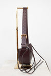 A dark burgundy leather shoulder bag with a wide adjustable strap and gold-tone buckle, displayed on a minimalist gold metal stand against a clean white background.