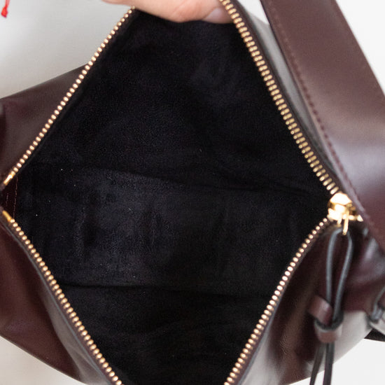 Close-up of a dark burgundy leather bag being opened by hands, showing a black fabric interior and gold-tone zipper hardware.