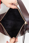 Close-up of a dark burgundy leather bag being opened by hands, showing a black fabric interior and gold-tone zipper hardware.