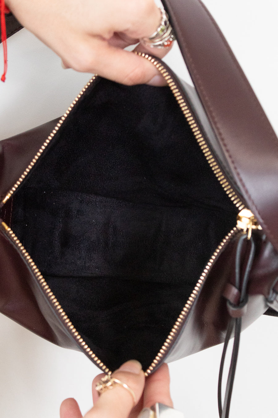 Close-up of a dark burgundy leather bag being opened by hands, showing a black fabric interior and gold-tone zipper hardware.