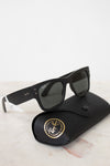 Black sunglasses with white accents on a black case with a gold logo, on a light marble surface.