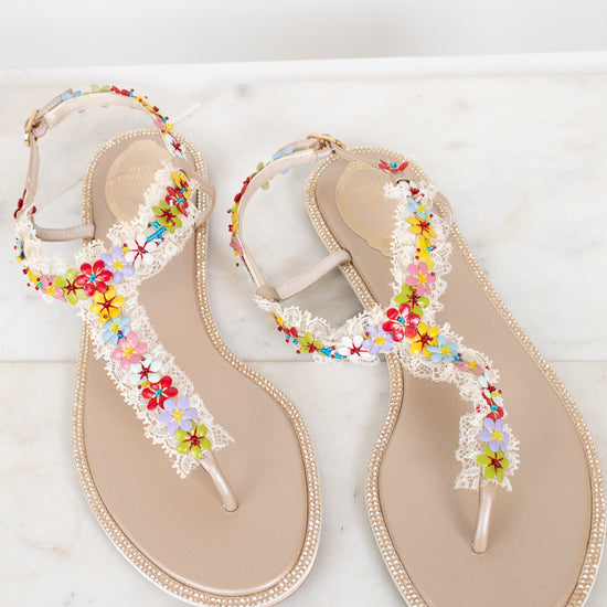 A pair of beige thong sandals decorated with white lace, rhinestone bordered soles, and colorful embroidered flowers, displayed on a light marble surface.