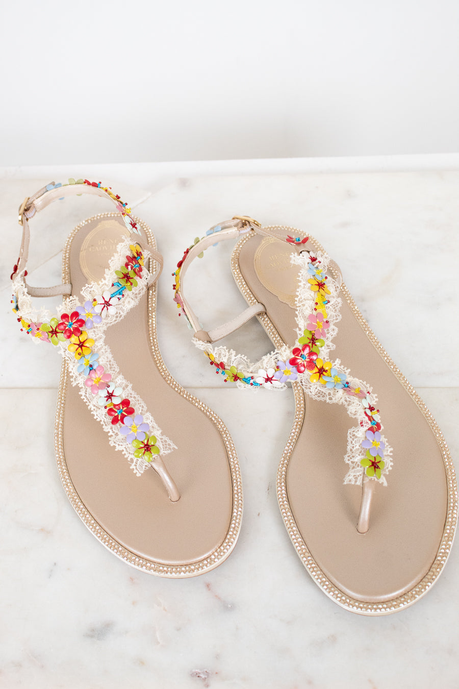 A pair of beige thong sandals decorated with white lace, rhinestone bordered soles, and colorful embroidered flowers, displayed on a light marble surface.