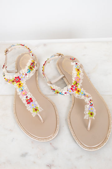 A pair of beige thong sandals decorated with white lace, rhinestone bordered soles, and colorful embroidered flowers, displayed on a light marble surface.