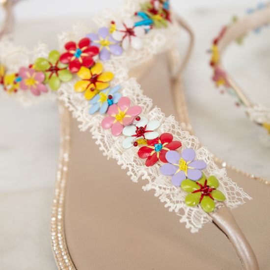 A close-up of a beige thong sandal decorated with white lace and colorful attached flowers set against a light neutral background.