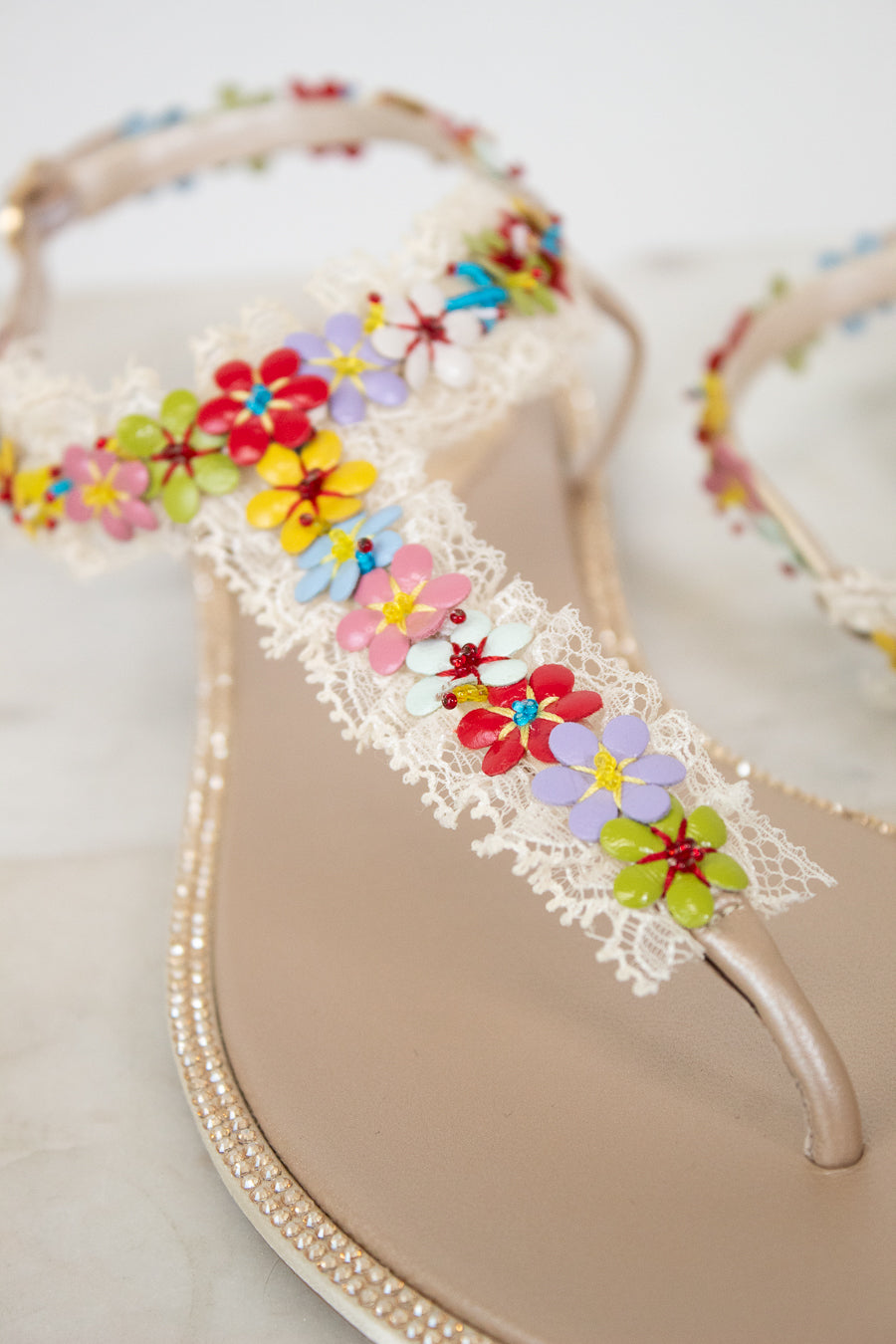 A close-up of a beige thong sandal decorated with white lace and colorful attached flowers set against a light neutral background.