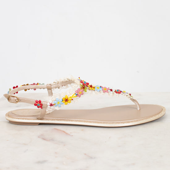 A single flat thong sandal with a beige footbed and a delicate floral strap embellished with colorful faux flowers resting on a light marble surface against a white background.