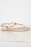 A single flat thong sandal with a beige footbed and a delicate floral strap embellished with colorful faux flowers resting on a light marble surface against a white background.