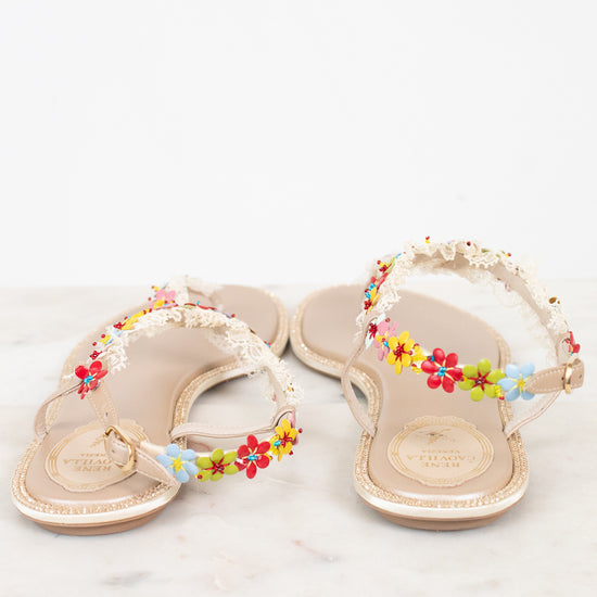 A pair of beige flat sandals decorated with colorful flowers and white lace detail on straps, displayed on a light marble surface against a white background.