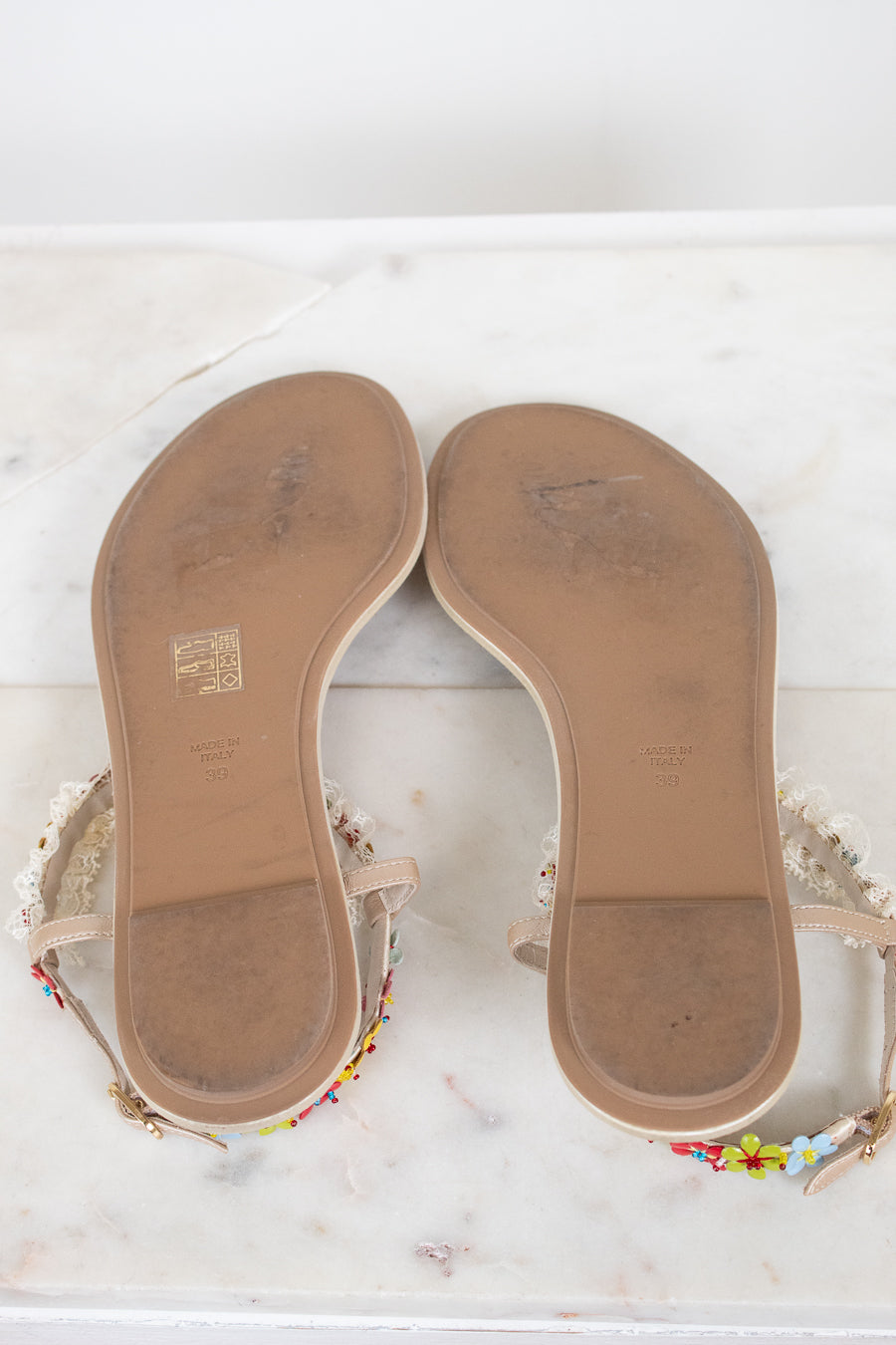 A pair of women's flat sandals shown from the bottom sole, resting on a white marble surface. 