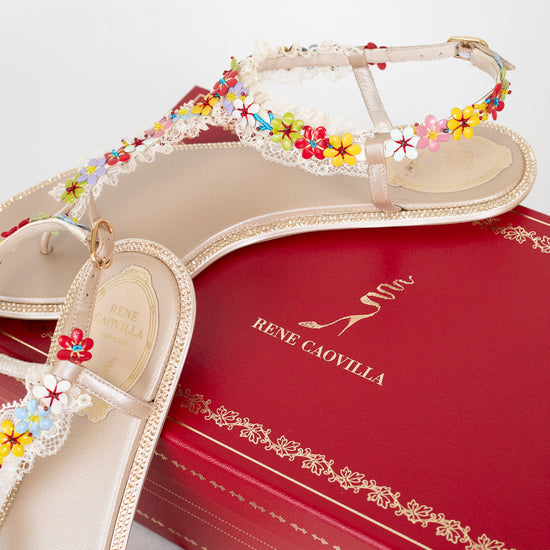 A pair of beige Rene Caovilla flat sandals decorated with colorful flowers and white lace trim, displayed on top of a red branded shoebox.