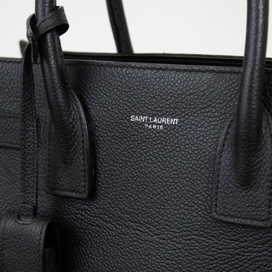 Close-up of a black pebbled leather Saint Laurent handbag with double top handles and a small silver-tone logo plaque on the front.