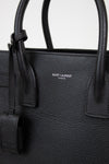 Close-up of a black pebbled leather Saint Laurent handbag with double top handles and a small silver-tone logo plaque on the front.