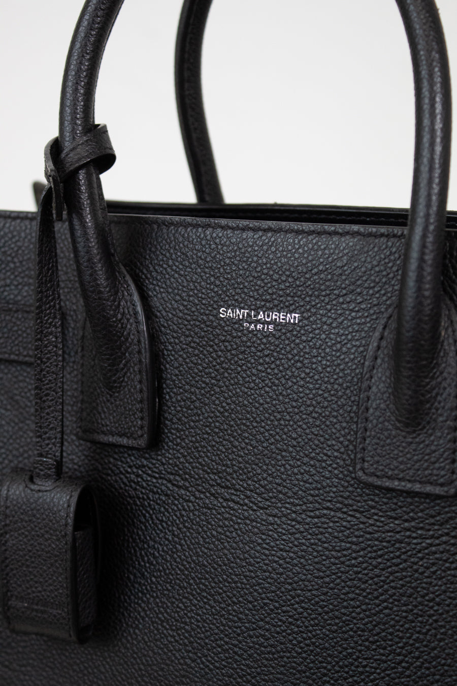 Close-up of a black pebbled leather Saint Laurent handbag with double top handles and a small silver-tone logo plaque on the front.