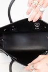 Close-up of the interior of a black leather handbag being held open by two hands wearing rings, set against a light marble background.