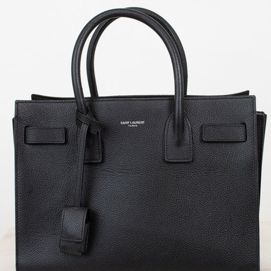 A black pebbled leather Saint Laurent handbag with double top handles, a hanging leather clochette, and a small silver-tone brand logo on the front, set against a light off-white background.