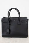 A black pebbled leather Saint Laurent handbag with double top handles, a hanging leather clochette, and a small silver-tone brand logo on the front, set against a light off-white background.