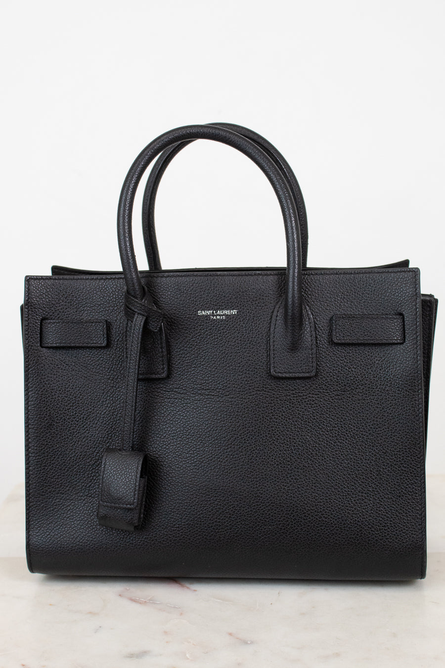 A black pebbled leather Saint Laurent handbag with double top handles, a hanging leather clochette, and a small silver-tone brand logo on the front, set against a light off-white background.