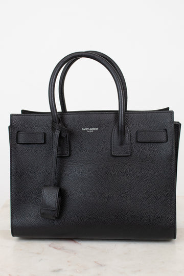 A black pebbled leather Saint Laurent handbag with double top handles, a hanging leather clochette, and a small silver-tone brand logo on the front, set against a light off-white background.