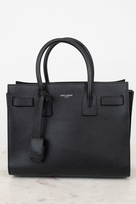 A black pebbled leather Saint Laurent handbag with double top handles, a hanging leather clochette, and a small silver-tone brand logo on the front, set against a light off-white background.