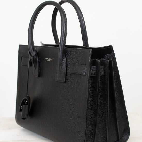A black pebbled leather handbag with double top handles, a front strap detail, and a hanging clochette, photographed on a light marble surface against a white background.