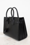 A black pebbled leather handbag with double top handles, a front strap detail, and a hanging clochette, photographed on a light marble surface against a white background.