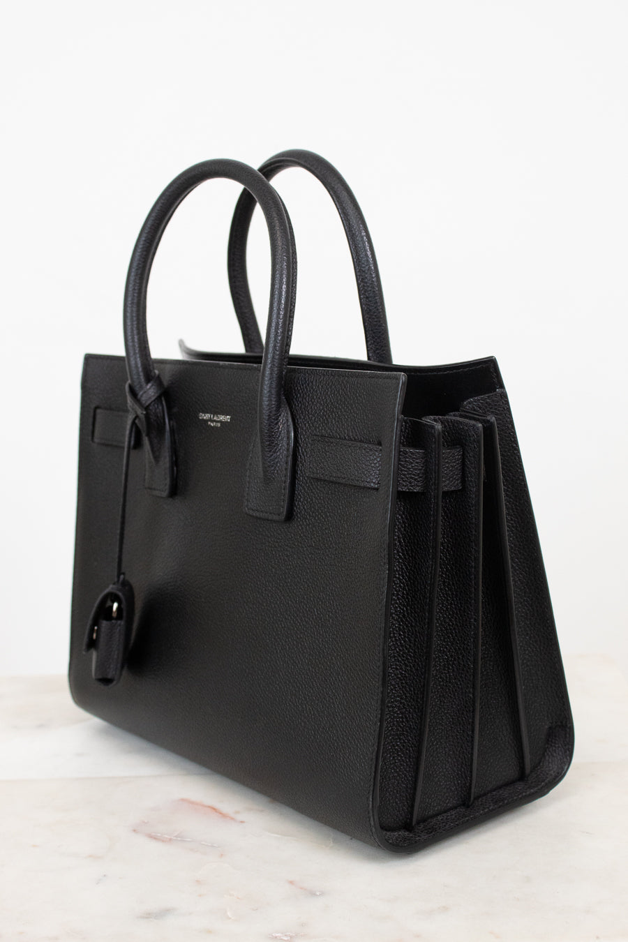 A black pebbled leather handbag with double top handles, a front strap detail, and a hanging clochette, photographed on a light marble surface against a white background.