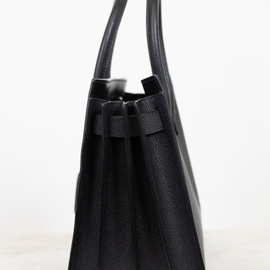 A black pebbled leather handbag shown from a side profile view, featuring dual top handles and a structured rectangular silhouette, set against a clean white background.