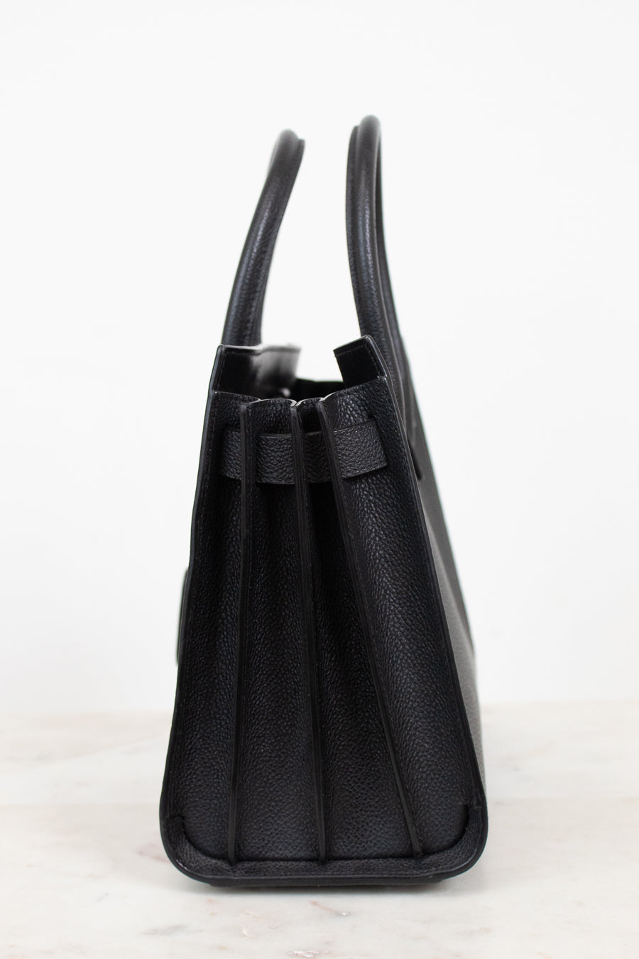 A black pebbled leather handbag shown from a side profile view, featuring dual top handles and a structured rectangular silhouette, set against a clean white background.