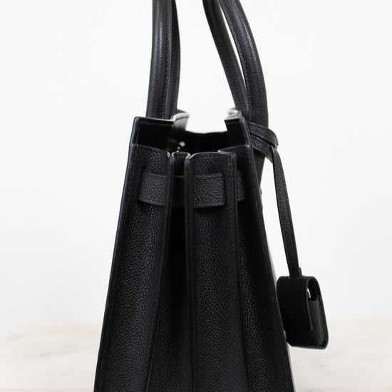 A black pebbled leather handbag shown from the side, featuring dual top handles, a front strap detail, and a hanging tag, set against a plain white background.