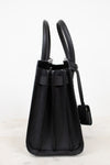 A black pebbled leather handbag shown from the side, featuring dual top handles, a front strap detail, and a hanging tag, set against a plain white background.