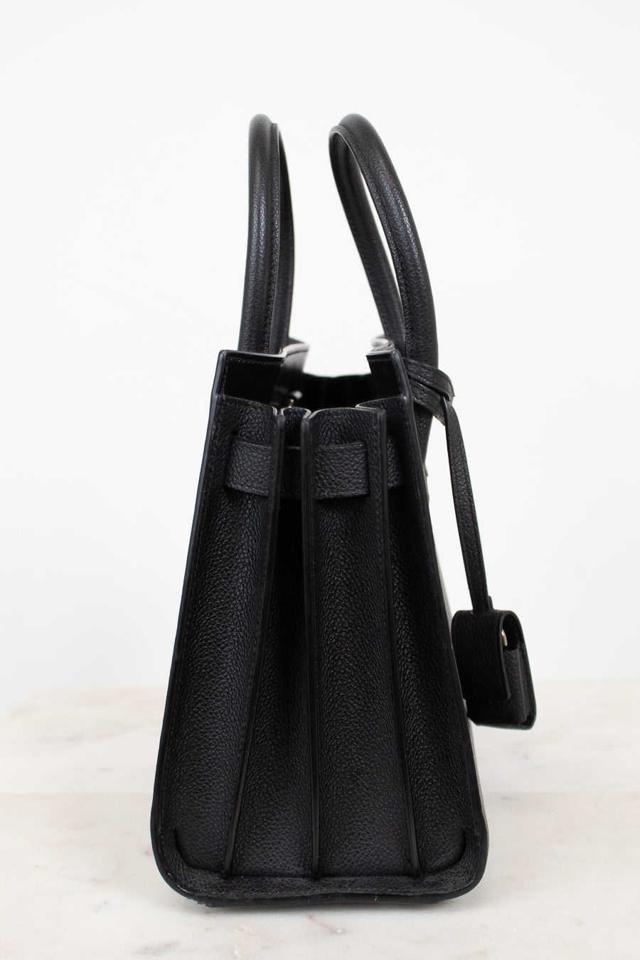 A black pebbled leather handbag shown from the side, featuring dual top handles, a front strap detail, and a hanging tag, set against a plain white background.