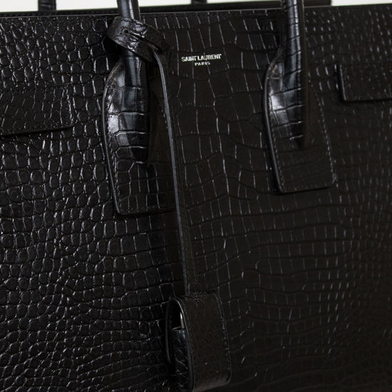 A close-up of a black crocodile-embossed leather handbag with dual top handles and a small gold-tone 'SAINT LAURENT PARIS' logo plaque on the front.
