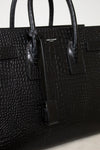 A close-up of a black crocodile-embossed leather handbag with dual top handles and a small gold-tone 'SAINT LAURENT PARIS' logo plaque on the front.