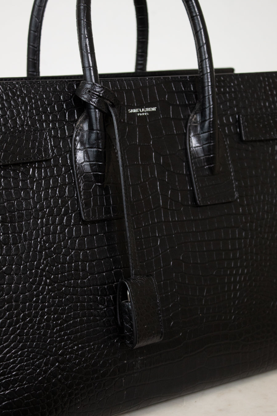 A close-up of a black crocodile-embossed leather handbag with dual top handles and a small gold-tone 'SAINT LAURENT PARIS' logo plaque on the front.