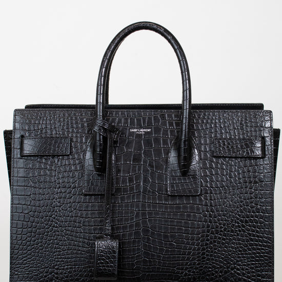 A black crocodile-embossed leather handbag with double top handles, a front flap detail, and a hanging clochette, photographed against a plain white background.