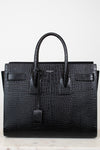 A black crocodile-embossed leather handbag with double top handles, a front flap detail, and a hanging clochette, photographed against a plain white background.