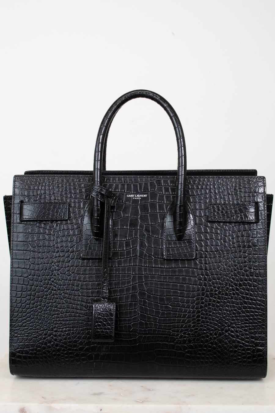 A black crocodile-embossed leather handbag with double top handles, a front flap detail, and a hanging clochette, photographed against a plain white background.