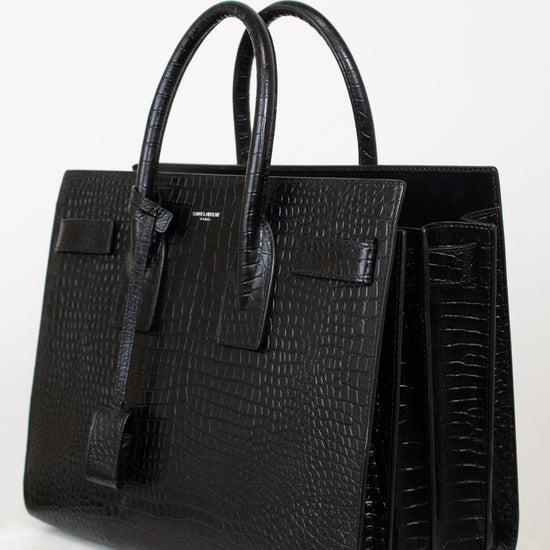 A black crocodile-embossed leather handbag with double top handles, displayed on a light marble surface against a white background.