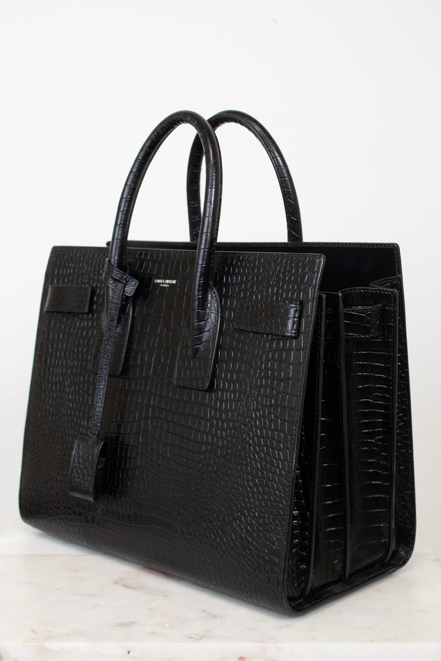 A black crocodile-embossed leather handbag with double top handles, displayed on a light marble surface against a white background.